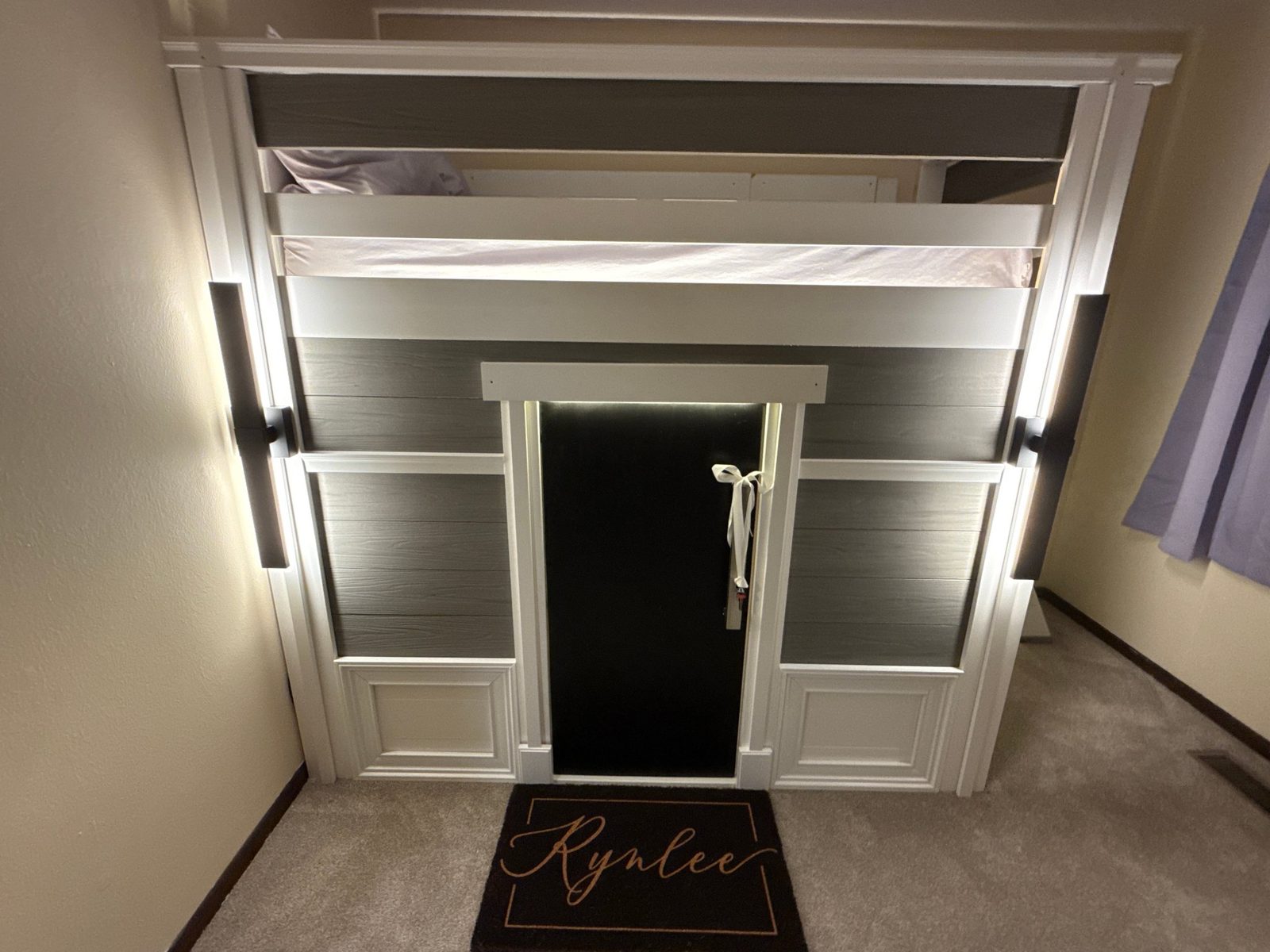 Front of the finished playhouse — gray cladding, white trim, black door, two lit sconces